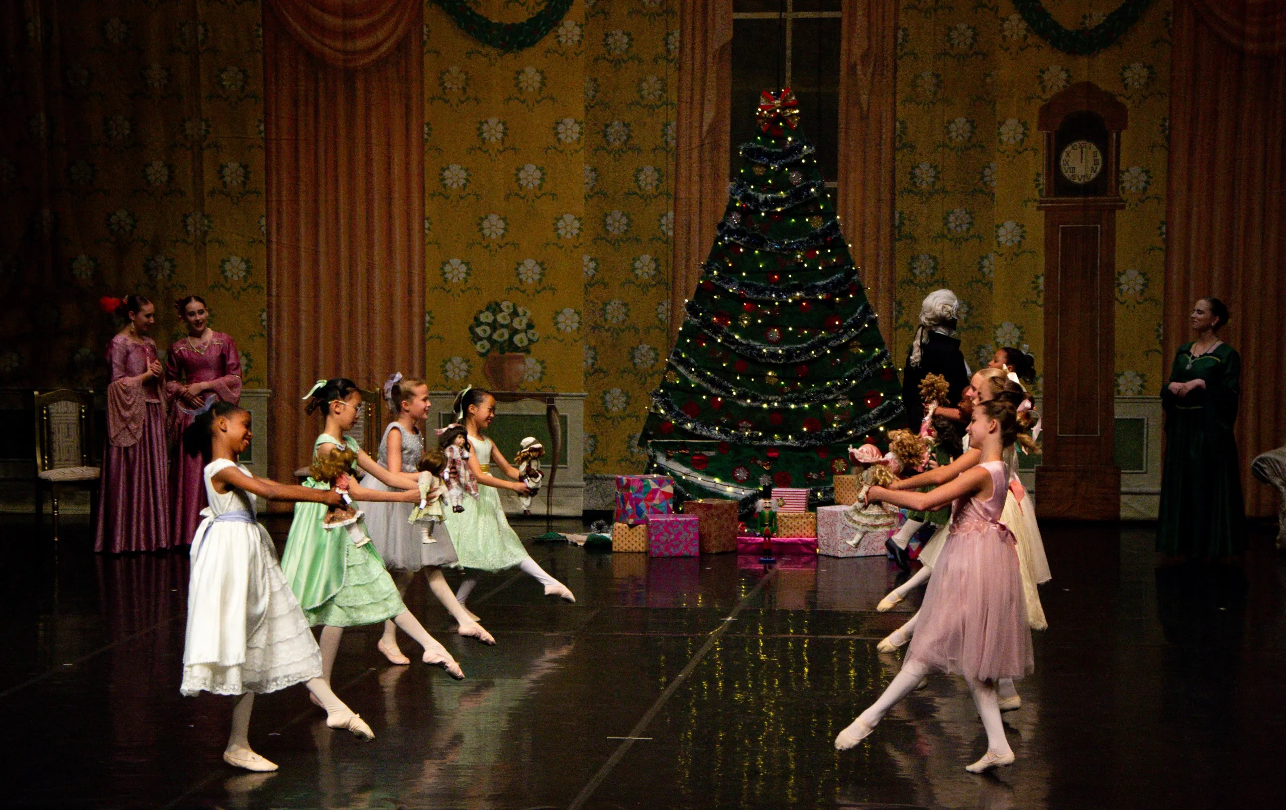 Wide group photograph from YRBC's 2023 performance of The Nutcracker ballet. Two groups of dancers face each other holding dolls with a Christmas tree in the background.